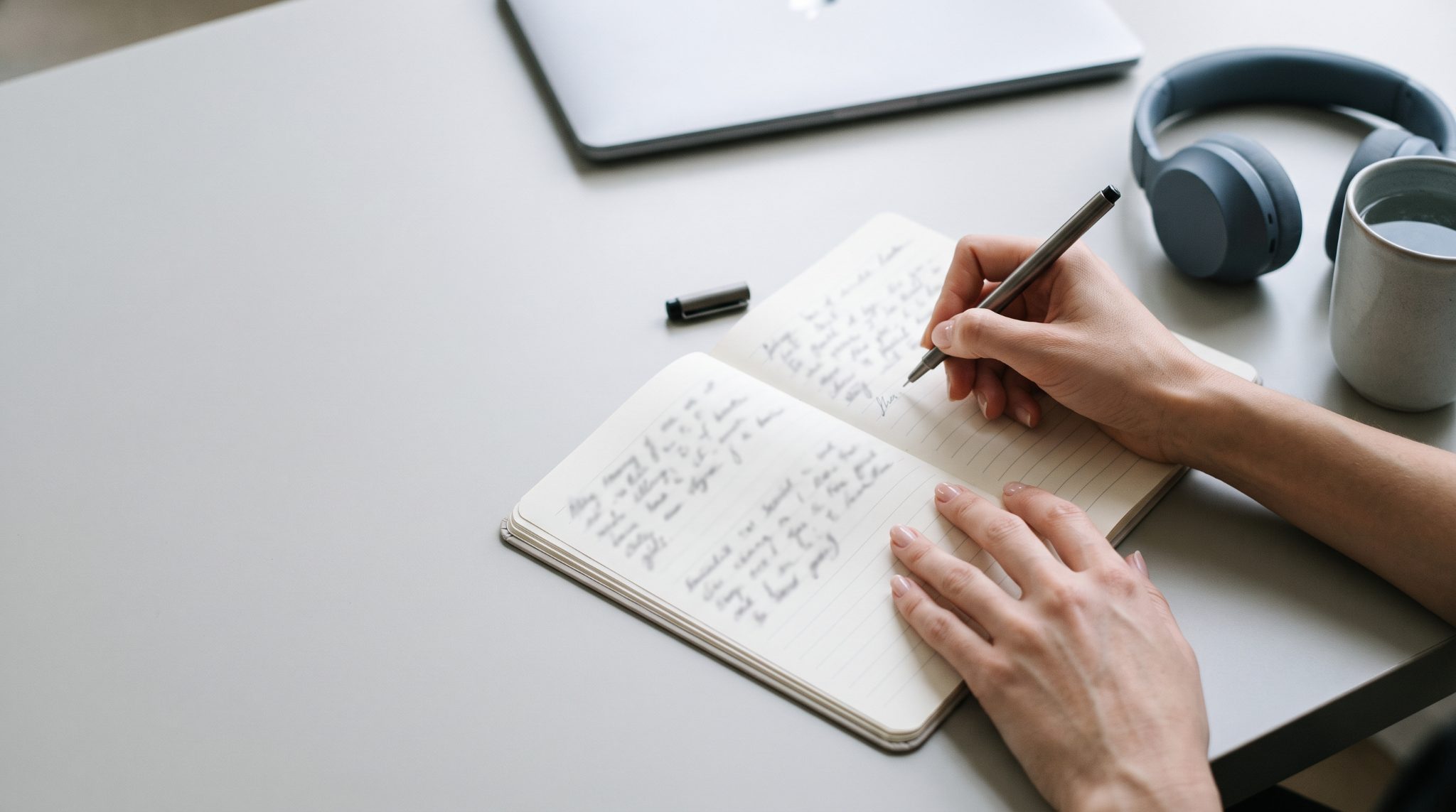 Hands writing in a notebook beside headphones on a clean silver-grey desk
